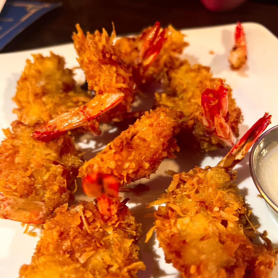 Crispy golden-brown coconut shrimp with red tails, served on a white plate with a dipping sauce.